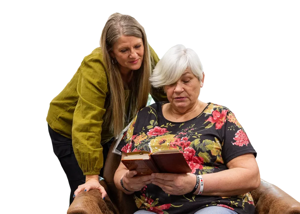 An older woman sits reading a book while another woman stands beside her, looking on with a gentle expression.