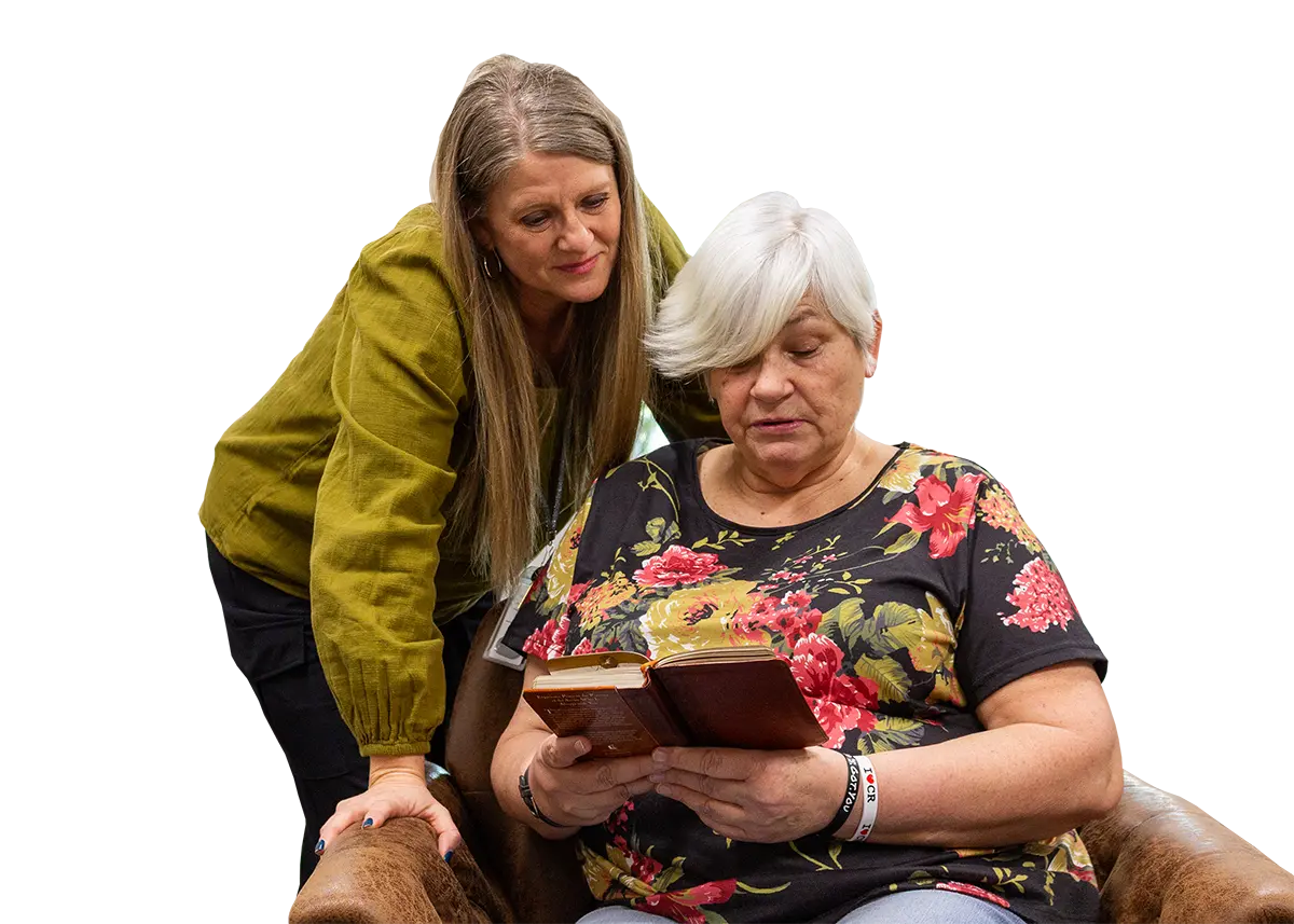 An older woman sits reading a book while another woman stands beside her, looking on with a gentle expression.
