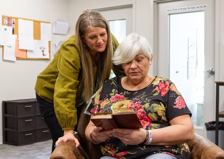 An older woman sits reading a book while another woman stands beside her, looking on with a gentle expression.