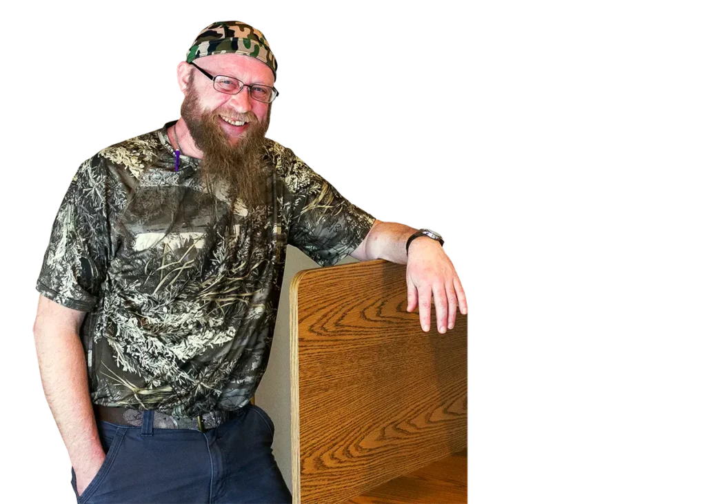 A smiling man in a camouflage shirt and head covering leans on a wooden desk divider.