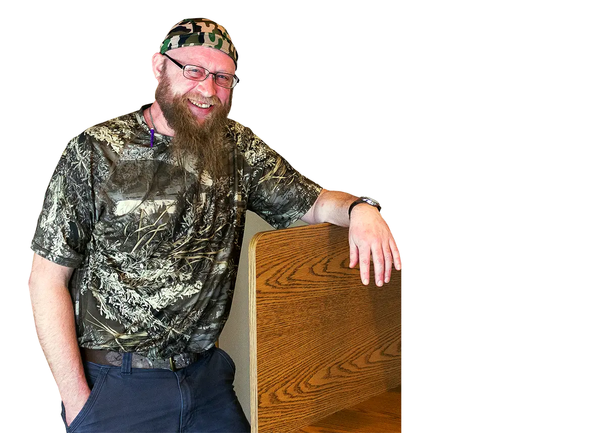 A smiling man in a camouflage shirt and head covering leans on a wooden desk divider.