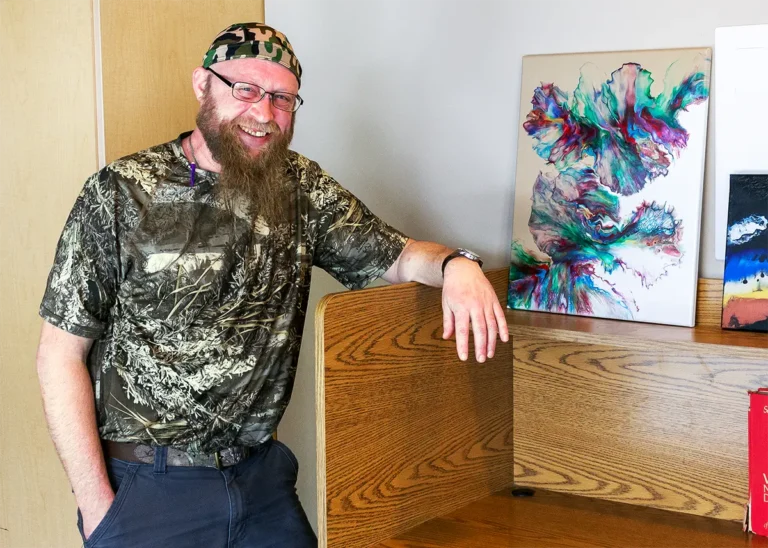 A smiling man in a camouflage shirt and head covering leans on a wooden desk divider, with an abstract painting in the background.